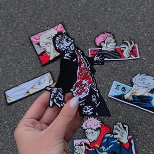 May include: A selection of embroidered anime character patches. The patches display different characters in action poses, with a central patch showing a figure in a black coat with red detailing. Other patches feature characters with pink hair and Japanese text.