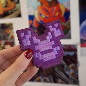 Pode incluir: Um patch de peitoral bordado roxo pixelizado. O patch tem formato quadrado com um recorte para o pescoço e cavas. O patch é segurado por uma mão com esmalte vermelho. Outras imagens estão no fundo.