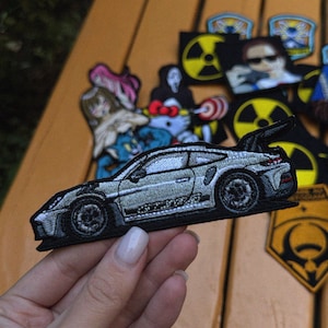 May include: A silver embroidered patch of a sports car with black accents. The patch is detailed with realistic shading and features a spoiler. Other patches with various designs are visible in the background.