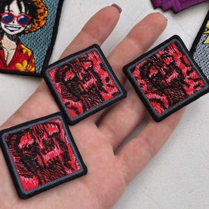Puede incluir: Parches bordados con un diseño rojo y negro dentro de un marco cuadrado con un borde gris. Los parches se sostienen en una mano, con otros parches visibles en el fondo, incluido un personaje de dibujos animados con un sombrero de paja y gafas de sol.