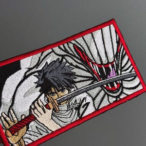 May include: Embroidered patch featuring an anime-style character with dark hair holding a sword. The design includes a red and white creature and is framed by a red border. The patch is rectangular.