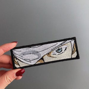 May include: Embroidered patch depicting an anime-style eye with a blue iris, partly concealed by a white bandage. The patch has a black border and is rectangular. The eye is set against a beige skin tone.