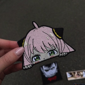 Op de afbeelding: Geborduurde patch met een rozeharige anime-personage met groene ogen en een zwarte omtrek. Het personage kijkt over een zwarte rand. De patch heeft een gedetailleerd ontwerp, voor kleding of accessoires.