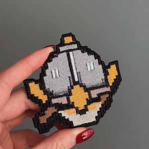 Può includere: Toppa ricamata con un design pixelato di una teiera. La toppa ha un bordo nero con dettagli grigi, gialli e marroni. Il design della teiera ricorda la grafica dei videogiochi retrò.