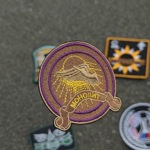 Könnte beinhalten: Gestickter Aufnäher mit einem braunen und lila kreisförmigen Design. Der Aufnäher zeigt einen stilisierten Vogel mit Sonnenstrahlen und das Wort "MOHOЛИТ" auf einem Banner. Weitere Aufnäher sind im Hintergrund zu sehen.
