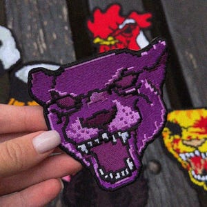 May include: A purple embroidered patch depicting a pixelated panther head with an open mouth and teeth. The patch has a black border and is held by a hand. Other patches with a red rooster and a yellow skull are visible in the background.