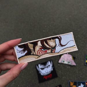 May include: Embroidered patch featuring a detailed anime character with brown hair and red markings on the face. The patch has a light blue background and a rectangular shape. Other anime patches are visible.