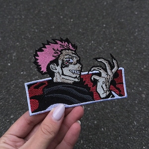 May include: Embroidered patch featuring a character with pink spiky hair, a sinister grin, and a skeletal hand. The patch is rectangular with a red and black background and a white border. The character is wearing a black outfit.