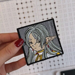 May include: Embroidered patch featuring an anime-style character with long, silver hair and pointed ears. The character has a serious expression and is wearing a white and gold outfit. The patch is square with a black border and is held by a hand with red nail polish.