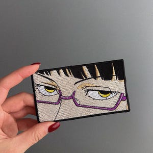May include: Embroidered patch featuring anime-style eyes with yellow irises, black eyelashes, and purple-framed glasses. The patch has a rectangular shape with a beige background and black hair.