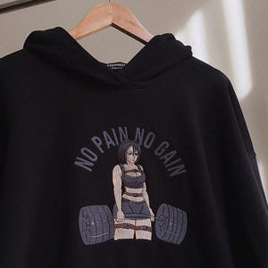以下が含まれることがあります： 「NO PAIN NO GAIN」の刺繍が施された黒いパーカー。女性がバーベルを持ち上げるイラストが描かれています。デザインは、トレーニングウェアを着た女性がバーベルを持ち上げている様子です。パーカーにはドローストリングフードが付いています。