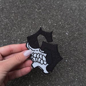 May include: A black and white embroidered patch in the shape of the letter "G" with a skull design. The patch is held by a hand with a light pink nail polish.
