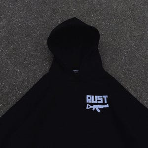 May include: Black hooded sweatshirt with the word "RUST" in a light blue, blocky font, and a light blue graphic of a rifle. The sweatshirt is made of a soft material and has a hood.