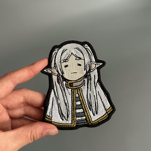 May include: Embroidered patch featuring a character with long white hair, pointed ears, and a white and gold trimmed coat. The patch has a black border and is held by a hand against a gray background. The character has a sad expression.