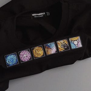 Op de afbeelding: Zwart T-shirt met ronde hals en zes geborduurde vierkante patches. Elke patch toont een uniek kleurrijk ontwerp. Het shirt heeft een label met de tekst "KEEPNEEDLE Made in Ukraine".