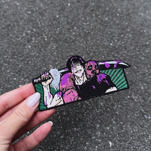 Puede incluir: Parche bordado con una ilustración estilo manga de dos personajes. Uno tiene cabello negro y un arma, el otro una máscara. El parche tiene un borde verde y detalles en morado y blanco.