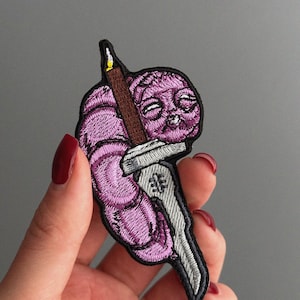 May include: Embroidered patch featuring a purple creature with a sword and a candle. The patch has a black border and is held by a hand with red nail polish. The creature has a smiling face and is holding a brown candle and a silver sword.
