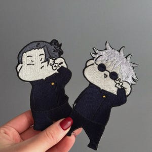Op de afbeelding: Twee geborduurde patches met stripfiguren in donkerblauwe uniformen. Het ene personage heeft zwart haar, het andere wit haar en een zonnebril. Beide patches hebben een kleine gouden knoop.