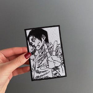 Puede incluir: Un parche rectangular en blanco y negro con una ilustración de estilo manga de una persona haciendo el signo de la paz. El parche tiene un borde negro y está sostenido por una mano con esmalte de uñas rojo.