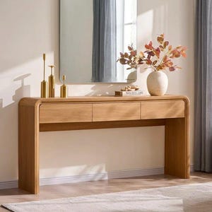 May include: A light oak wood console table with three drawers and rounded edges. On top are three gold-toned metal candle holders, a book, and a beige ceramic vase containing a branch with reddish-brown leaves, positioned below a rectangular mirror.