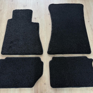 Puede incluir: Juego de cuatro alfombrillas negras para coche. Las alfombrillas son rectangulares con una superficie texturizada y un borde oscuro. Dos alfombrillas son más grandes, para la parte delantera, y dos más pequeñas, para la trasera.