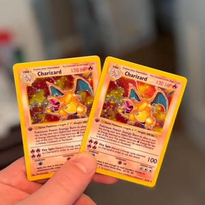 Op de afbeelding: Twee identieke Charizard-verzamelkaarten met een holografische illustratie van het oranje, draakachtige wezen met blauwe vleugels. De kaarten tonen 120 HP, de classificatie als Vlam-Pokémon en de tekst voor de Energy Burn en Fire Spin aanvallen.