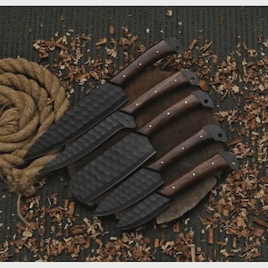 May include: A set of six black kitchen knives with textured blades and dark wooden handles. The knives are arranged on a wooden cutting board, surrounded by wood shavings and a coiled rope, creating a rustic aesthetic.