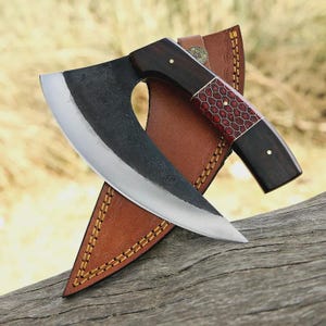 May include: A handcrafted cleaver knife with a dark wooden handle and a red honeycomb pattern. The blade is curved and has a silver edge. The knife rests in a brown leather sheath with decorative stitching.
