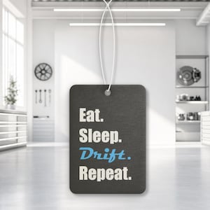 May include: A rectangular black air freshener hanging from a white string. The text "Eat. Sleep. Drift. Repeat." is printed on the front in a bold, sans-serif font. The word "Drift." is highlighted in a light blue color, while the others are white.