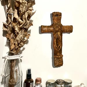 May include: A rustic, wooden cross with a carved design hangs on a white wall. A bouquet of dried roses sits in a glass bottle. Various jars and bottles are arranged on a wooden shelf below.