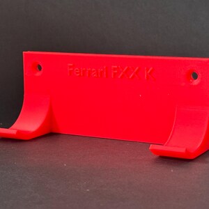 May include: A bright red wall mount with the text "Ferrari FXX K" printed on it. The mount has two curved support arms and two holes for mounting. The object is made of plastic.