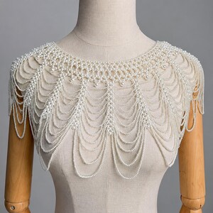 May include: A shoulder-covering capelet constructed from multiple strands of small white faux pearls. The design features a dense, woven collar section that transitions into cascading, scalloped layers of draped pearl chains that hang over the shoulders.