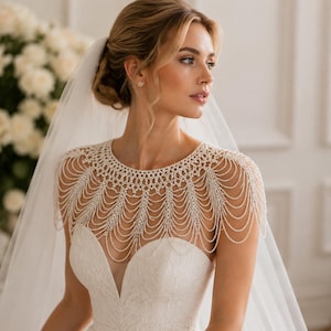 May include: A white bridal shoulder necklace featuring intricate layers of draped pearl-like beads and small clear crystals. The accessory covers the shoulders and upper chest, featuring a scalloped edge design that hangs over a strapless white dress.