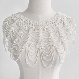 May include: A white pearl beaded shoulder necklace featuring a woven collar design with multiple cascading draped strands of small white pearls. The accessory covers the upper chest and shoulders, creating a layered, scalloped fringe effect.