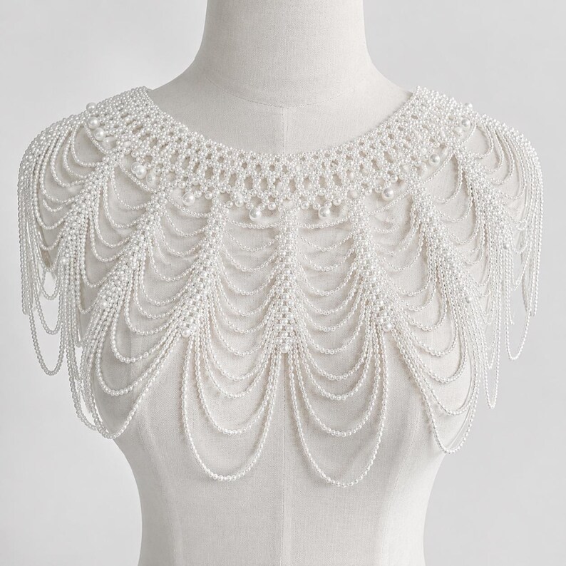 May include: A white pearl beaded shoulder necklace featuring a woven collar design with multiple cascading draped strands of small white pearls. The accessory covers the upper chest and shoulders, creating a layered, scalloped fringe effect.