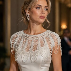 May include: A white satin dress featuring an intricate, multi-layered pearl beaded capelet overlay. The capelet consists of numerous draped strands of small white pearls that extend over the shoulders and across the chest in a scalloped pattern.