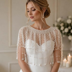 May include: A white pearl beaded capelet featuring multiple draped strands of small round pearls and teardrop-shaped pearl accents. The accessory covers the shoulders and upper chest, creating a layered fringe effect over a plain white garment.
