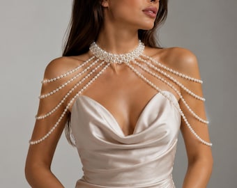 May include: A white pearl body chain featuring a high-neck choker collar connected to multiple draped strands of pearls that extend across the shoulders and down the upper arms, worn over a sleeveless cream-colored satin dress with a cowl neckline.