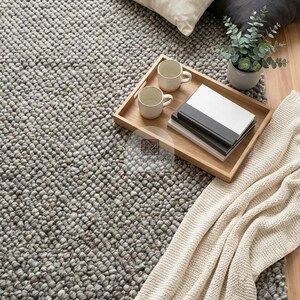 May include: A gray textured wool rug with a chunky bobble weave. On the rug sits a rectangular wooden tray holding two beige ceramic mugs and a stack of two books. Nearby are a cream pillow, a dark gray pillow, a small potted plant, and a cream throw.