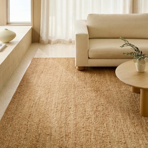 May include: A rectangular, woven jute rug in a warm, natural tan color. The rug is placed on a light-colored floor in a living room setting, with a beige sofa and round wooden coffee table visible.