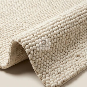 May include: A cream-colored area rug with a thick, textured surface made of small, rounded, pebble-like tufts. The rug is folded over to reveal a woven backing and features a small, square logo with the text KALEENRUGSTORE in the center.