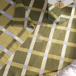 Puede incluir: Una alfombra tejida con un patrón de cuadros en verde oliva, crema, azul claro y amarillo. La alfombra es rectangular con una esquina doblada. Una mesa auxiliar hexagonal con espejo y una lámpara son visibles en el fondo.