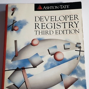 May include: A book cover with a white background and an abstract design featuring a gray crescent shape, red circles, and blue and gray squares. The title "Developer Registry Third Edition" is printed in black on the cover. The book is published by Ashton-Tate.