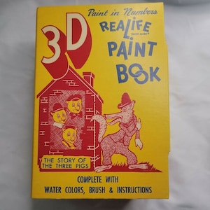 Op de afbeelding: Een gele doos met een rood 3D-schilderboek met het verhaal van de drie kleine biggetjes. De doos bevat waterverf, een kwast en instructies. Op de voorkant van de doos staat "Paint in Numbers Realife Paint Book".