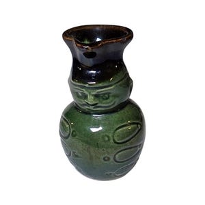 May include: A green ceramic pitcher shaped like a stylized figure with a dark brown glazed rim and spout. The body features embossed oval details and a molded face with defined eyes, nose, and mouth, finished with a glossy, reflective surface.