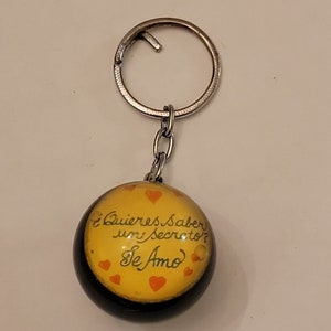 May include: A yellow and black keychain with a round charm that says "¿Quieres saber un secreto? Te Amo" with red hearts.