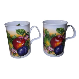 May include: Two white ceramic mugs featuring a colorful botanical illustration of red apples, dark purple plums, green leaves, and small pink flowers against a soft yellow background. Each mug has a curved handle and a smooth, cylindrical body.