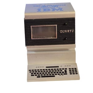 May include: A silver and black desk clock shaped like a vintage computer. The top features the text WMA JA VOLUNTEER 1983-1984 presented by IBM. The black screen area includes the word QUARTZ. A silver keyboard base is attached at the bottom.