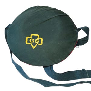 May include: A dark green Girl Scout canteen cover with a yellow and gold Girl Scout logo. The canteen cover has a round shape with a strap and a patterned interior. The canteen cover is made of fabric.