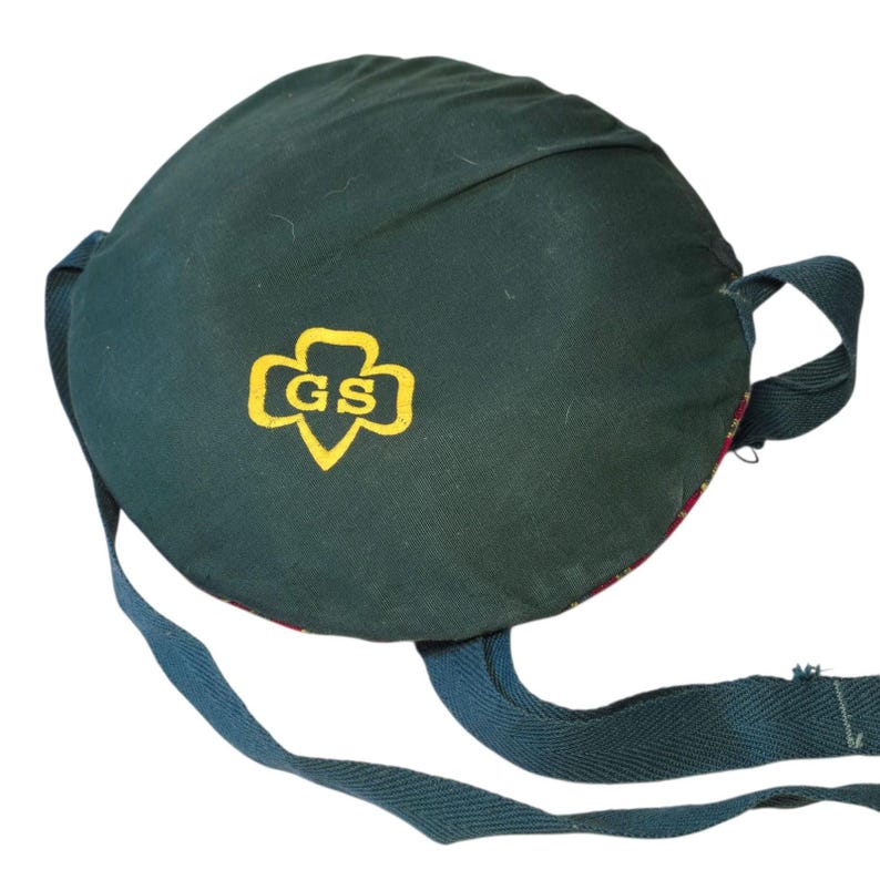 May include: A dark green Girl Scout canteen cover with a yellow and gold Girl Scout logo. The canteen cover has a round shape with a strap and a patterned interior. The canteen cover is made of fabric.